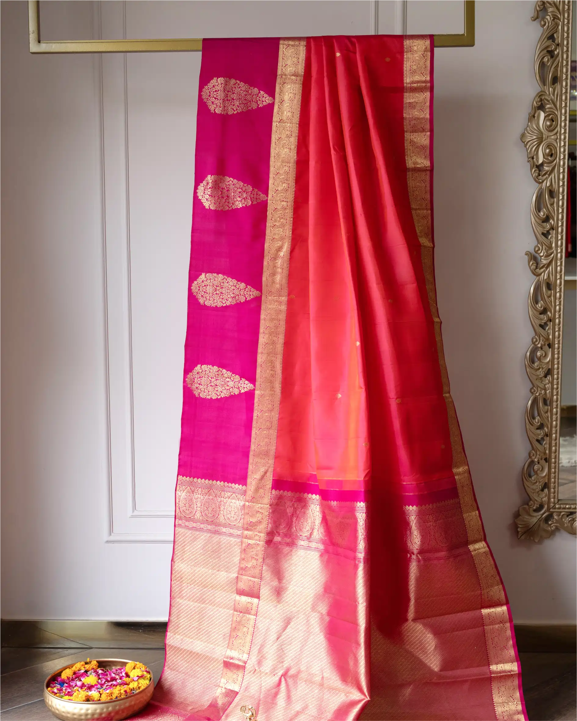 Handwoven red and pink Kanjivaram silk saree with a traditional gold zari border and buta motifs, displayed on a gold-framed hanger.