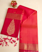 A folded and draped pink and red silk saree with a gold floral pattern on the pallu and ornate gold borders, displayed on a light cream background with scattered rose petals and a small gold decorative object.