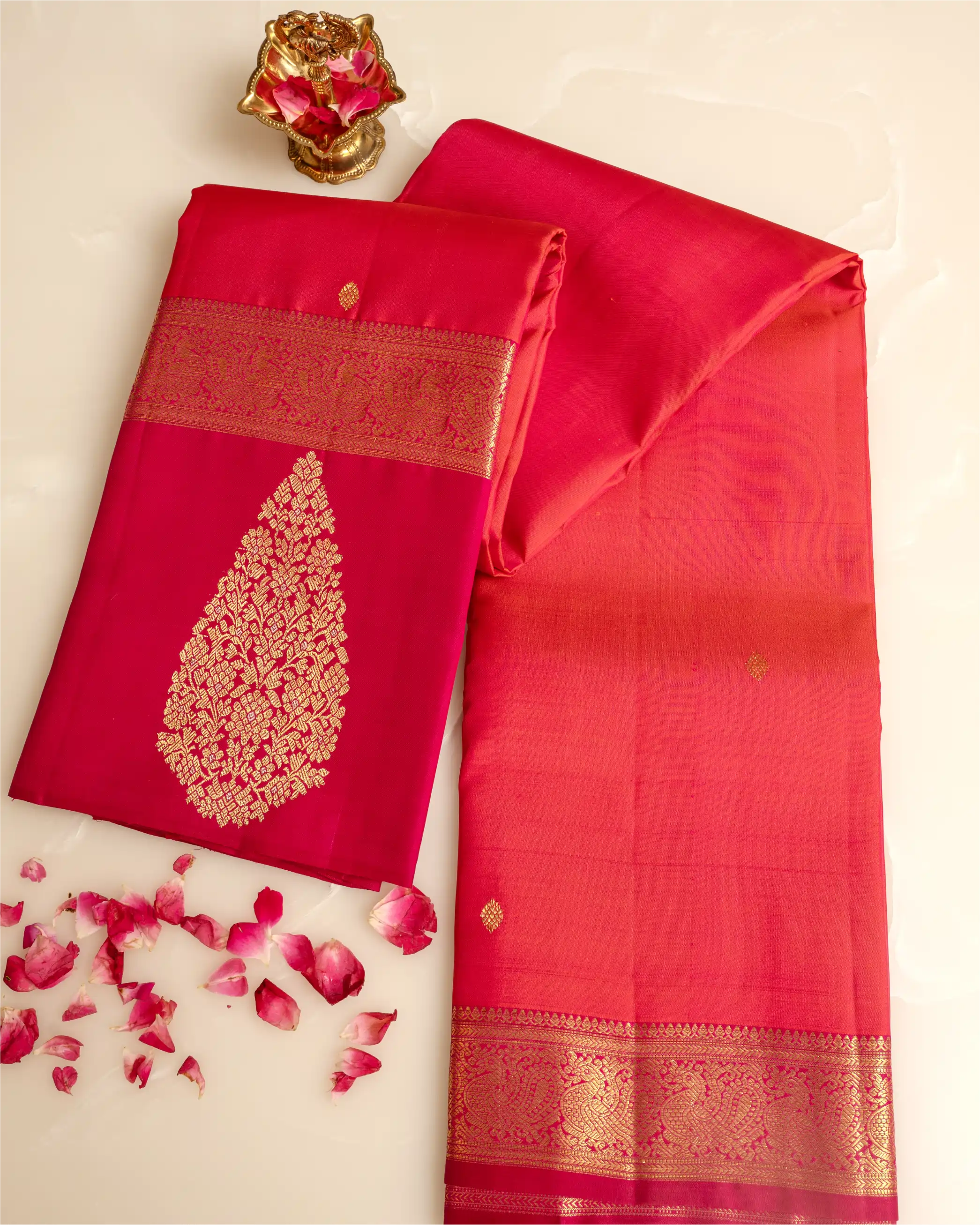 A folded and draped pink and red silk saree with a gold floral pattern on the pallu and ornate gold borders, displayed on a light cream background with scattered rose petals and a small gold decorative object.