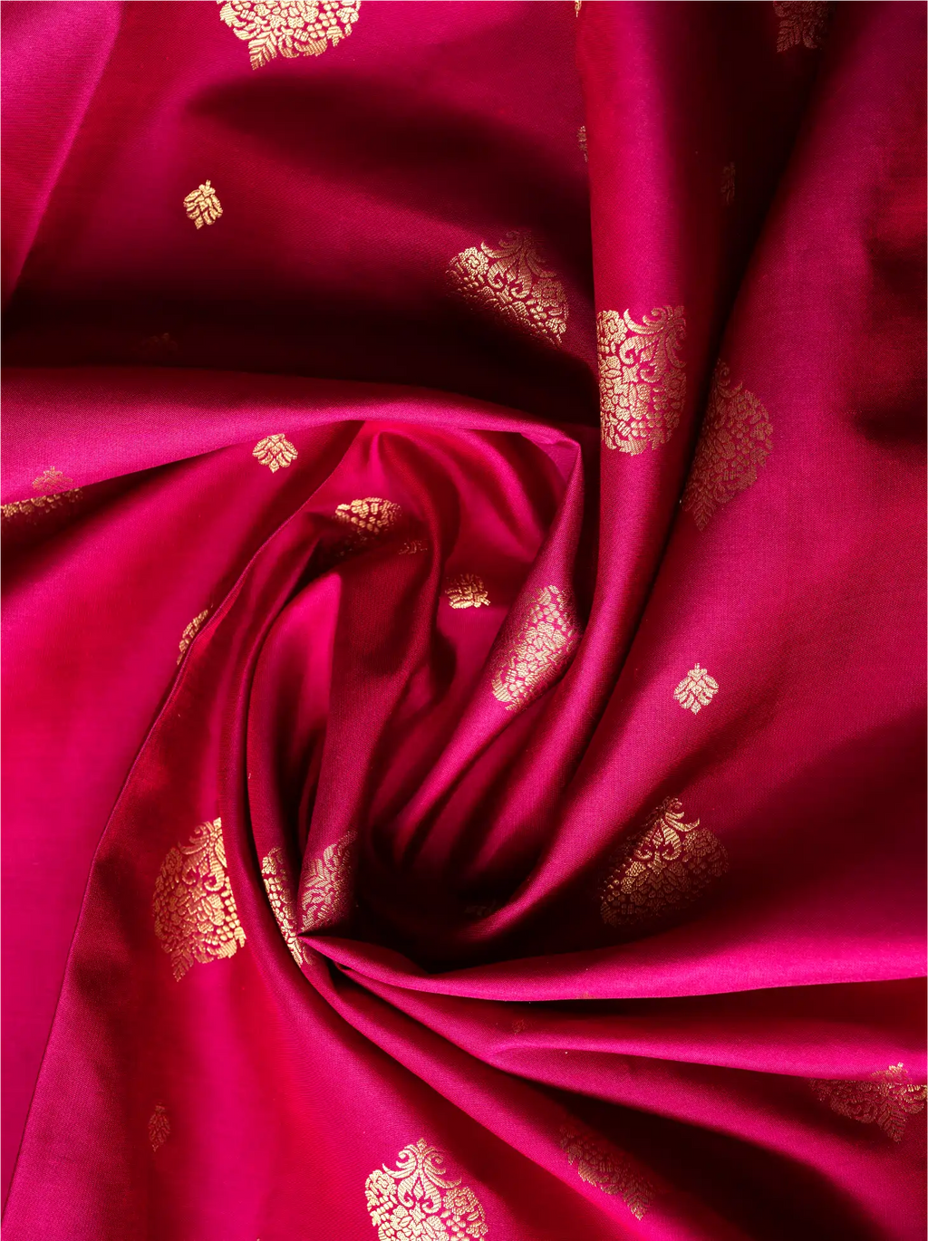 Close-up view of swirled vibrant magenta-red pure silk saree fabric showcasing small, intricate gold zari motifs woven across the glossy surface.