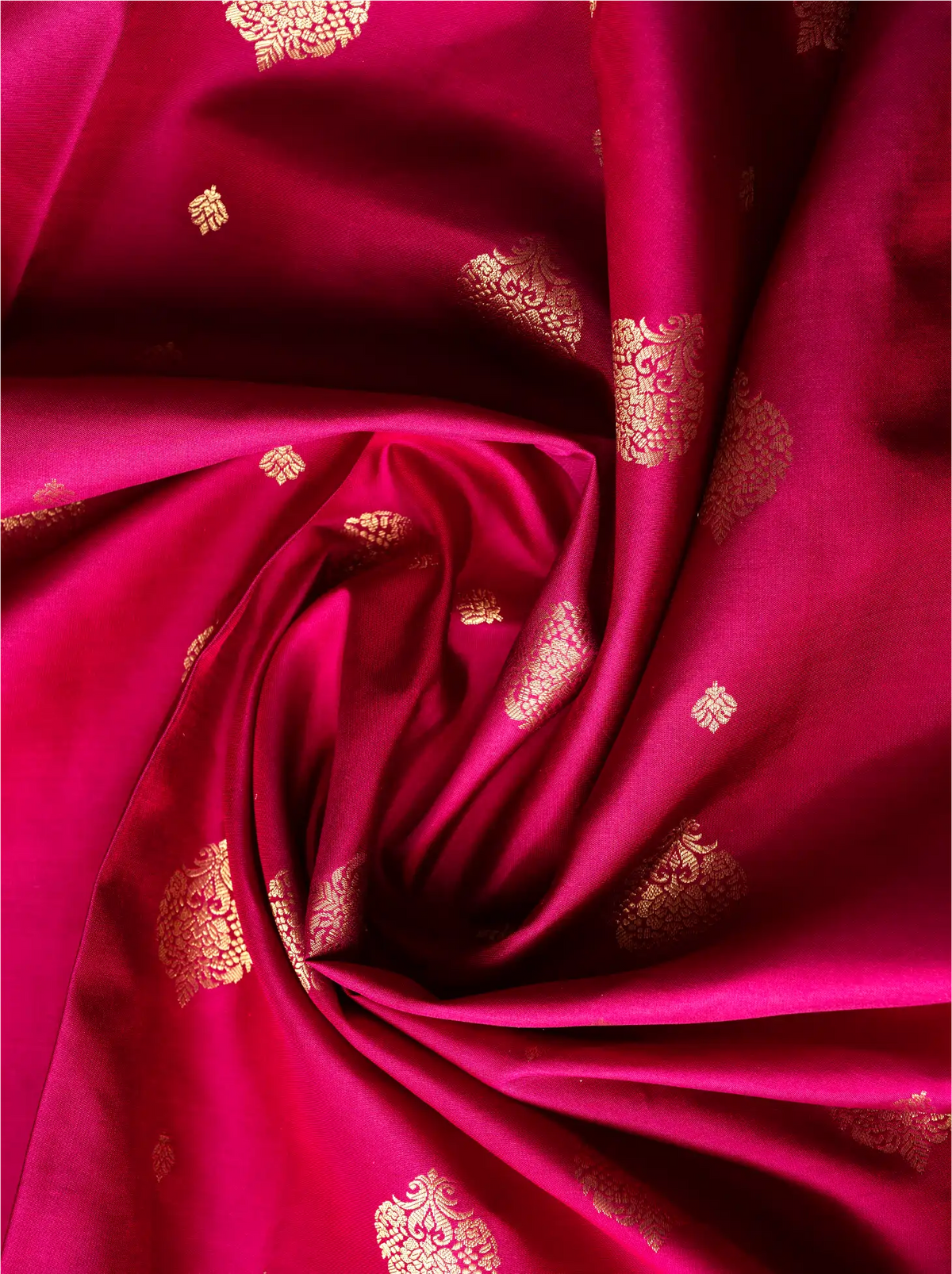 Close-up view of swirled vibrant magenta-red pure silk saree fabric showcasing small, intricate gold zari motifs woven across the glossy surface.