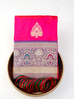 Folded red pure Katan silk saree with gold zari motifs displayed in a brass bowl with green and red glass bangles.