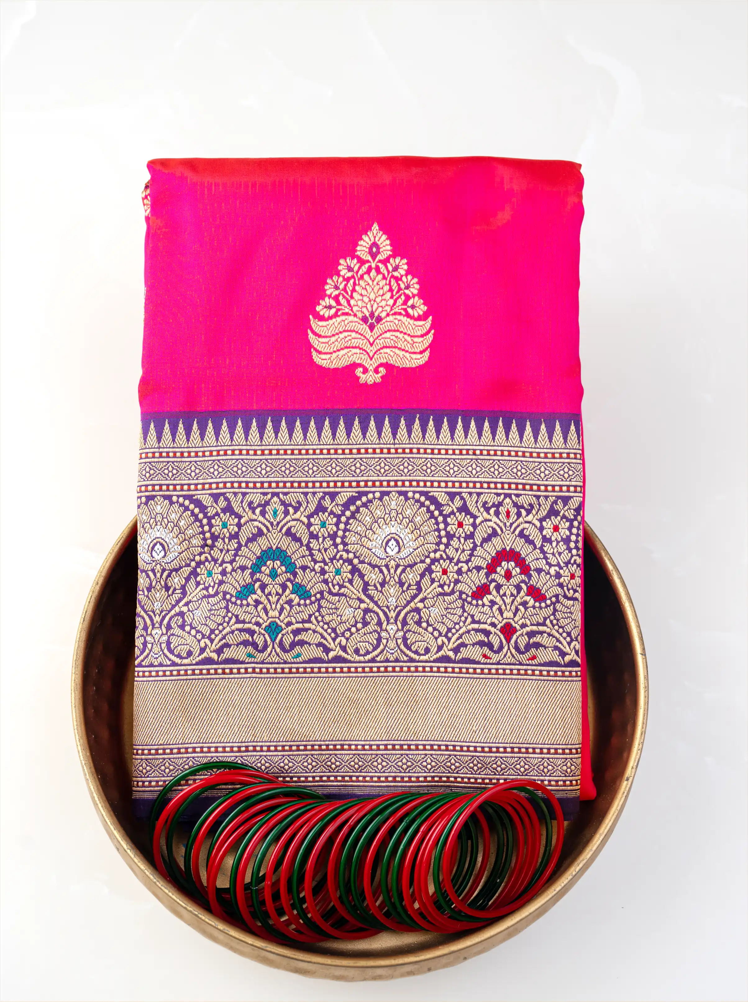Folded red pure Katan silk saree with gold zari motifs displayed in a brass bowl with green and red glass bangles.