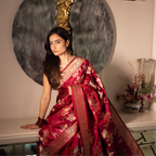 Model elegantly seated indoors wearing a premium Red Handwoven Katan Silk Saree with intricate gold zari floral motifs and a rich traditional border.