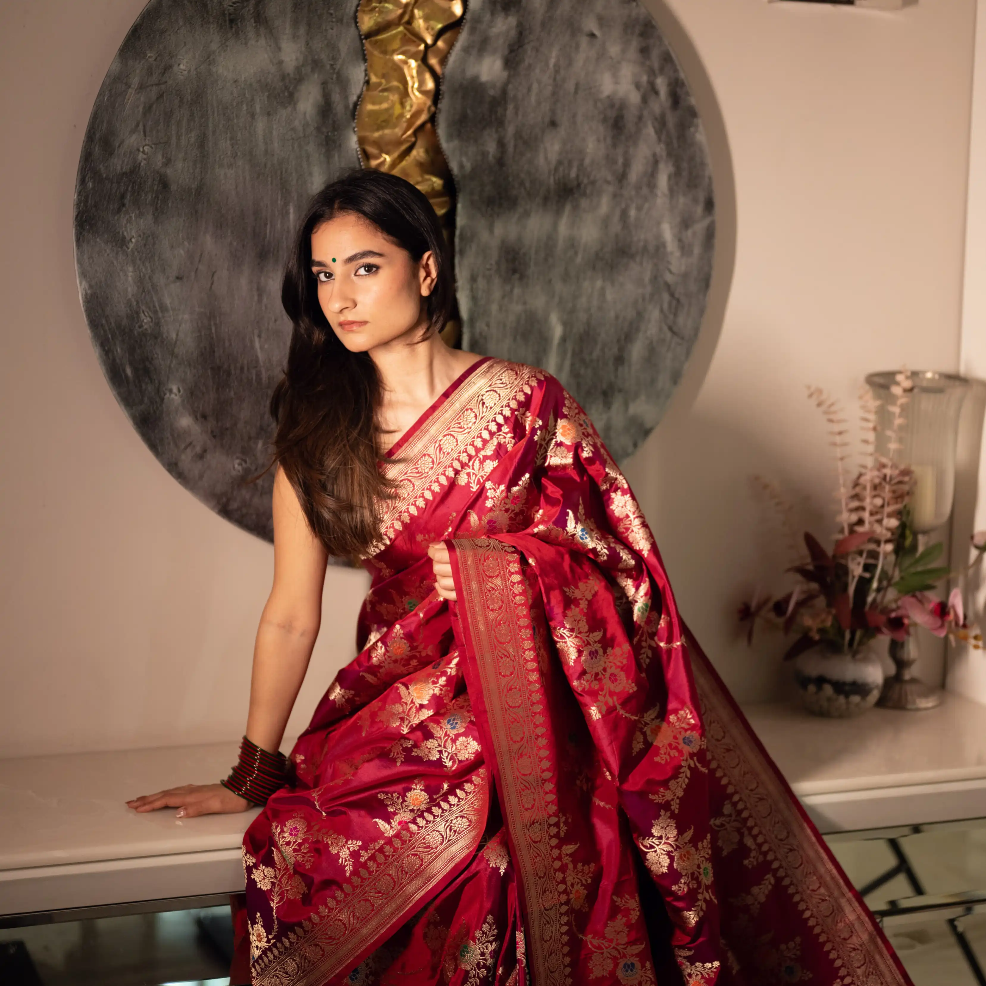 Model elegantly seated indoors wearing a premium Red Handwoven Katan Silk Saree with intricate gold zari floral motifs and a rich traditional border.