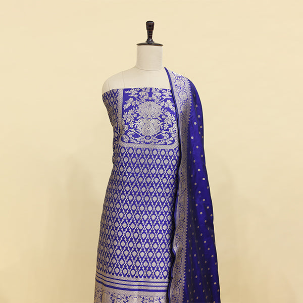 A mannequin displaying an elegant royal blue Banarasi silk suit featuring intricate silver zari work, traditional floral motifs, and a matching draped dupatta.
