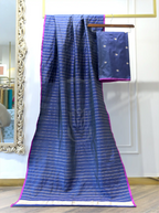 A bright blue silk saree with thin gold stripes and a wide gold border, draped over a hanger with its matching pink blouse piece hanging beside it in an indoor room.