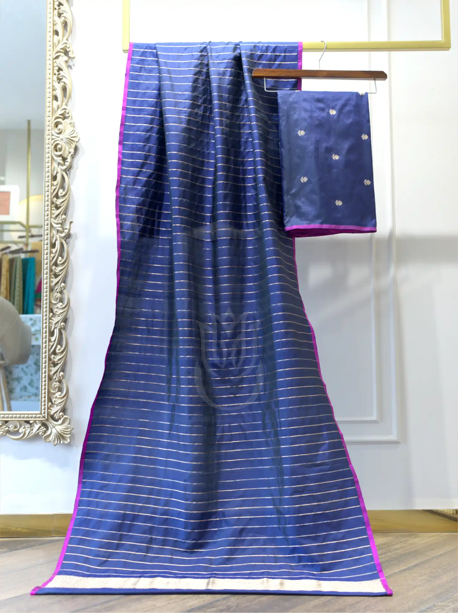A bright blue silk saree with thin gold stripes and a wide gold border, draped over a hanger with its matching pink blouse piece hanging beside it in an indoor room.