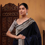 A woman wearing a royal blue Mashru silk saree with an abstract Jamdaani weave pattern and a silver border, standing next to a carved wooden screen.