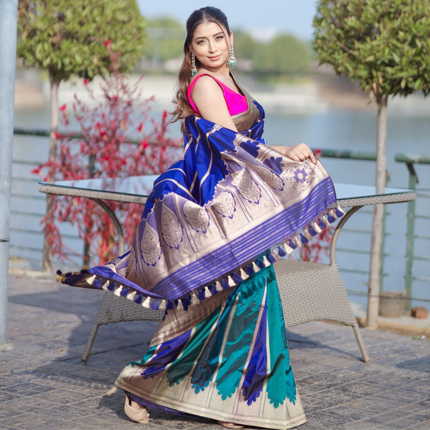 A model wearing a premium royal blue and teal silk saree with a wide gold border and tassel detailing, paired with a vibrant pink blouse.