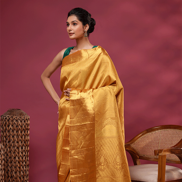 A model showcasing the Royal Mustard Kanjivaram silk saree with premium gold Suchi Zari work, styled with a contrasting sleeveless emerald green blouse against a deep red backdrop.