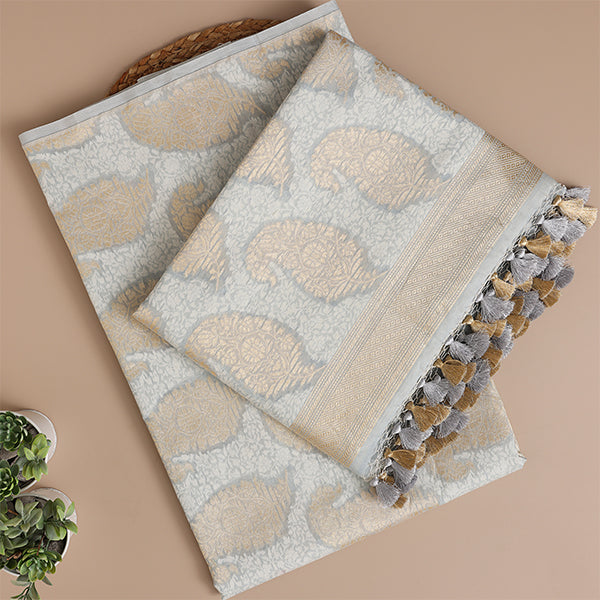 A flat-lay view of a luxurious silver-grey Banarasi silk suit fabric featuring intricate gold paisley motifs, a soft woven texture, and elegant tassel detailing on the dupatta edges.