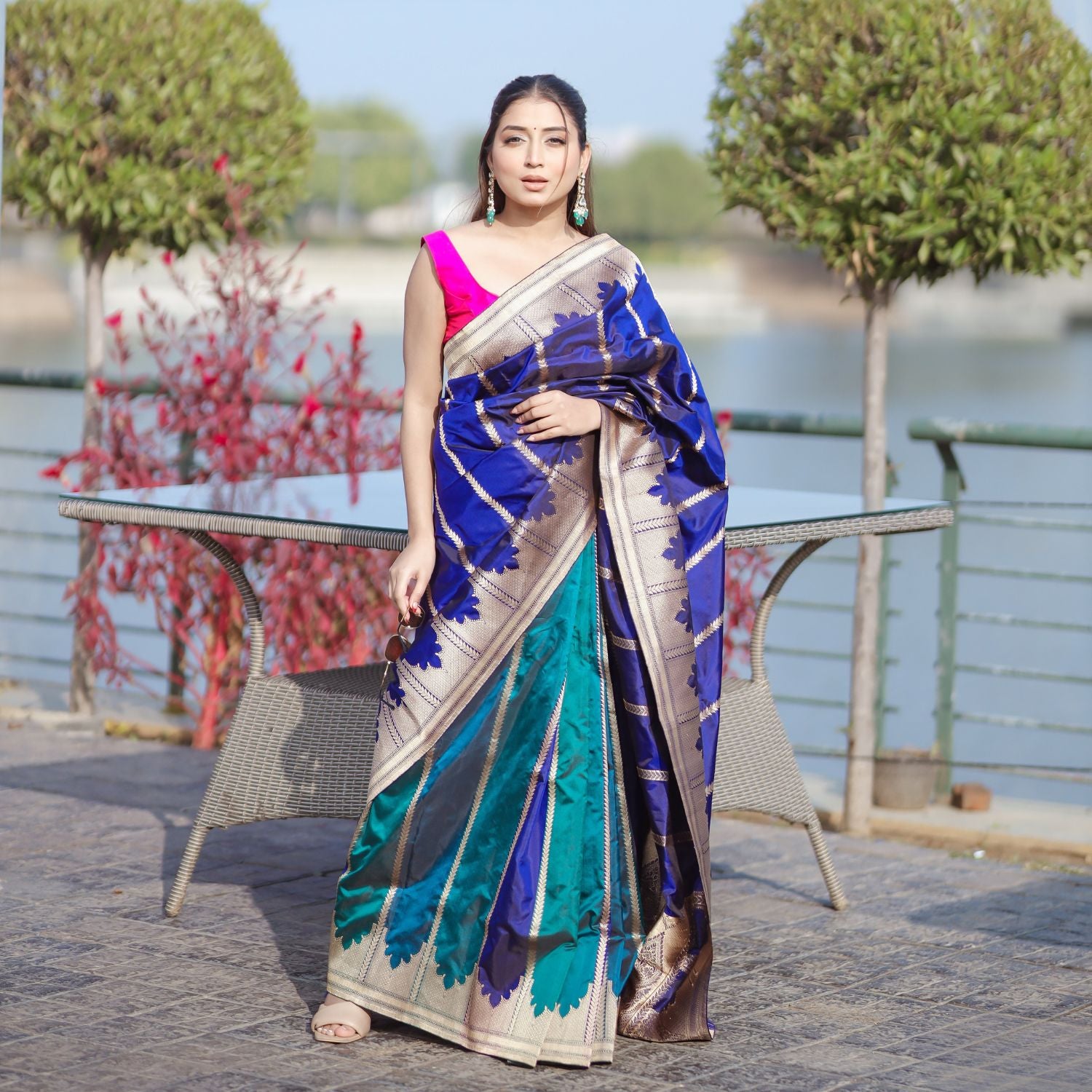 A model wearing a premium teal and royal blue silk saree with a wide gold border, styled with traditional jewelry in an outdoor setting.