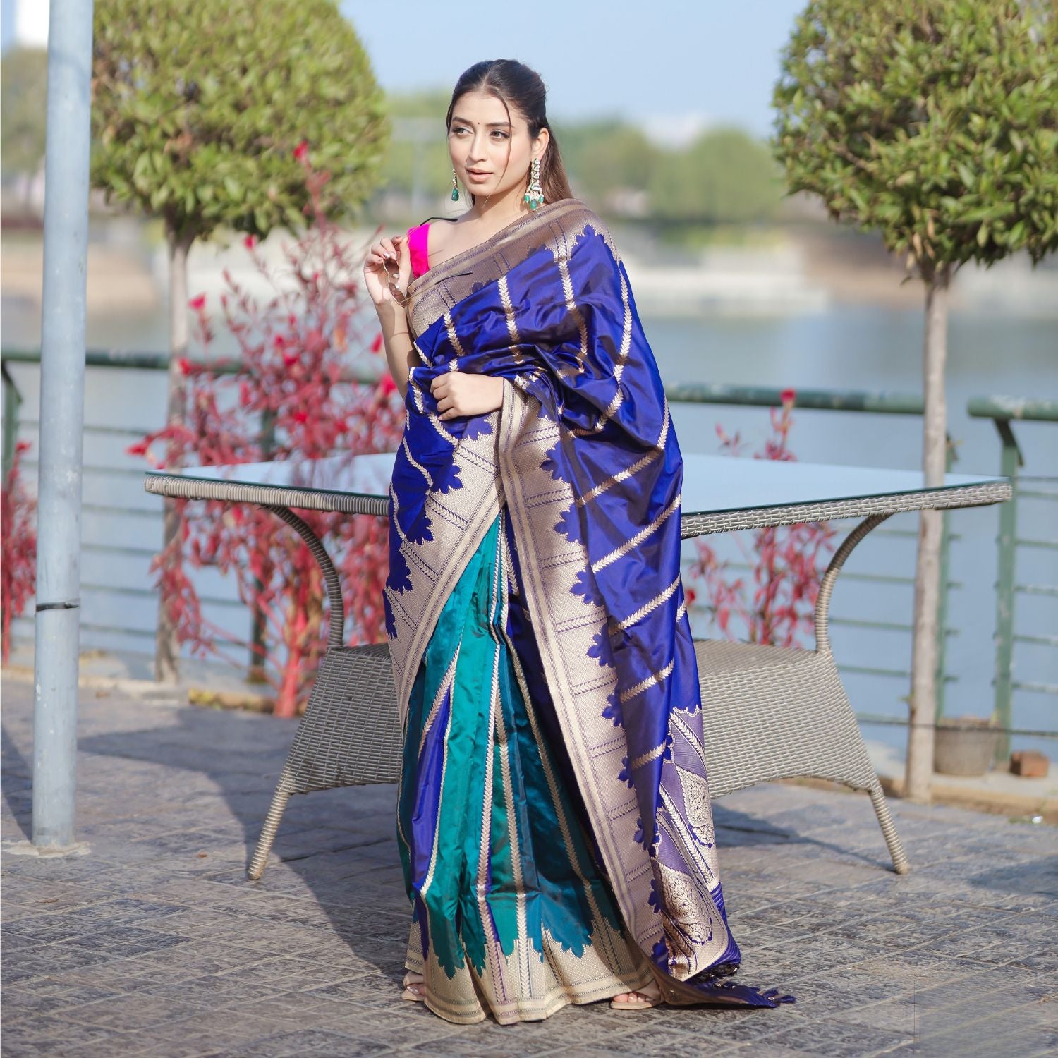 A model wearing a premium teal and royal blue silk saree with a wide gold border, styled with traditional jewelry in an outdoor setting.