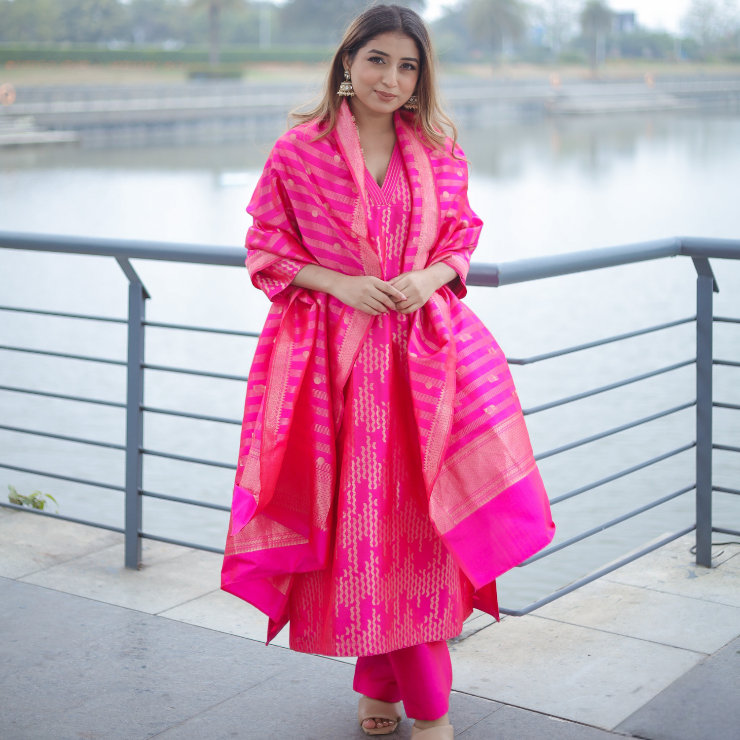 A model posing outdoors near a waterfront bridge, wearing a vibrant pink handwoven Banarasi silk suit with gold zari embroidery and a matching striped organza dupatta.