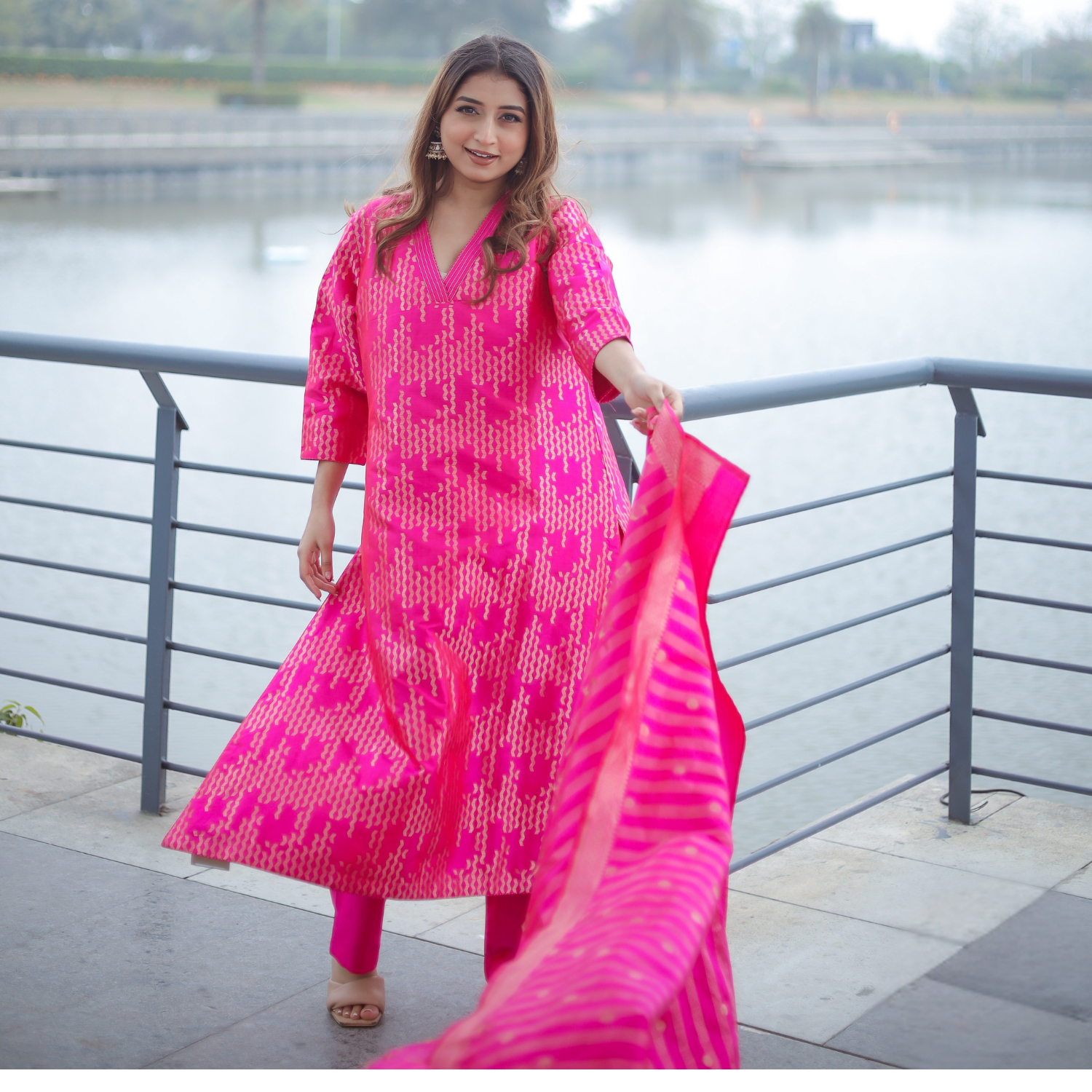 A model posing outdoors by a railing, wearing a vibrant pink handwoven Banarasi silk suit with gold zari embroidery and a matching striped organza dupatta.