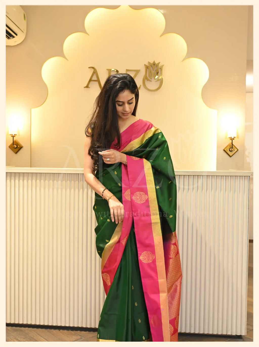 Front view of a woman wearing a handwoven dark green Mysore silk saree with a pink zari border standing inside an Arzo store.