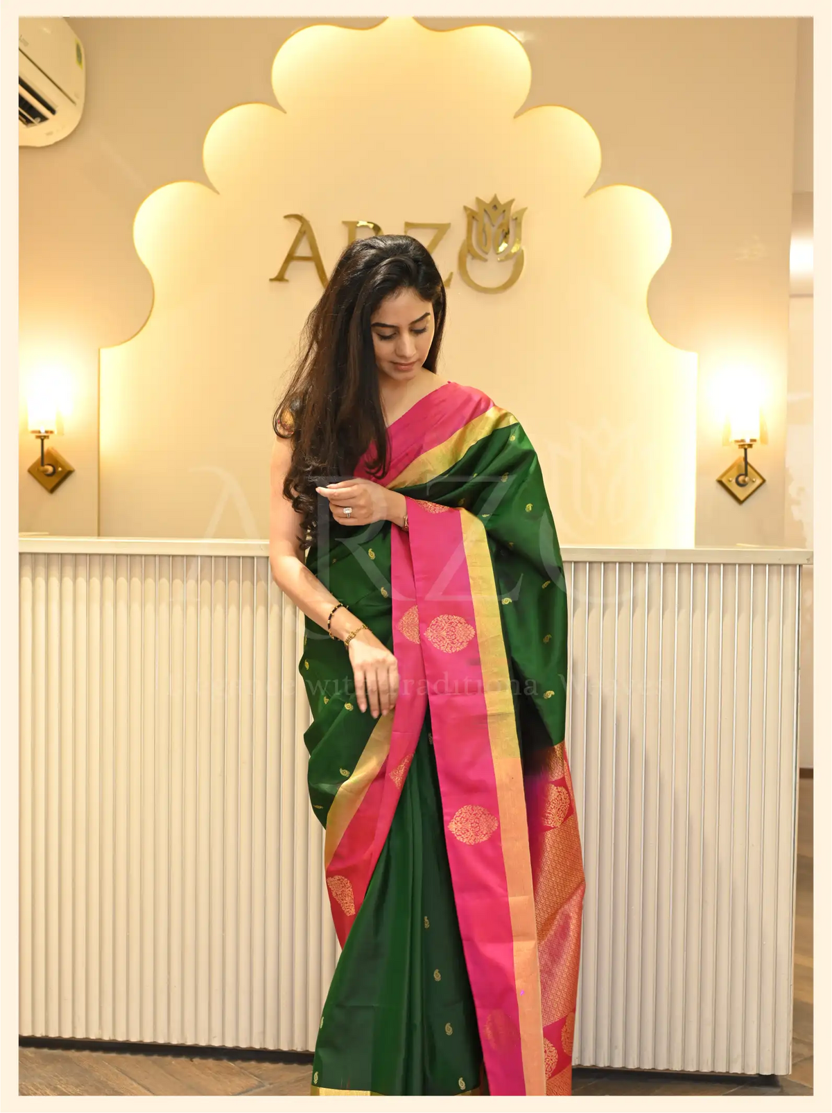 Front view of a woman wearing a handwoven dark green Mysore silk saree with a pink zari border standing inside an Arzo store.